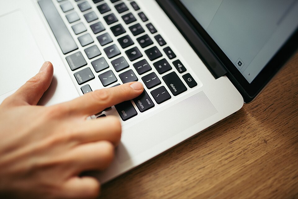 A hand is pressing keys on a laptop keyboard. The sleek device is positioned on a wooden desk, suggesting a focused work or study environmen